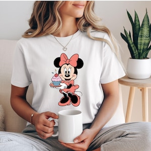 Puede incluir: Camiseta blanca con un dise&ntilde;o de dibujos animados de Minnie Mouse. Minnie est&aacute; representada con un vestido y lazo rosa, sosteniendo una magdalena en un plato. Ilustraci&oacute;n colorida para fans de Disney.