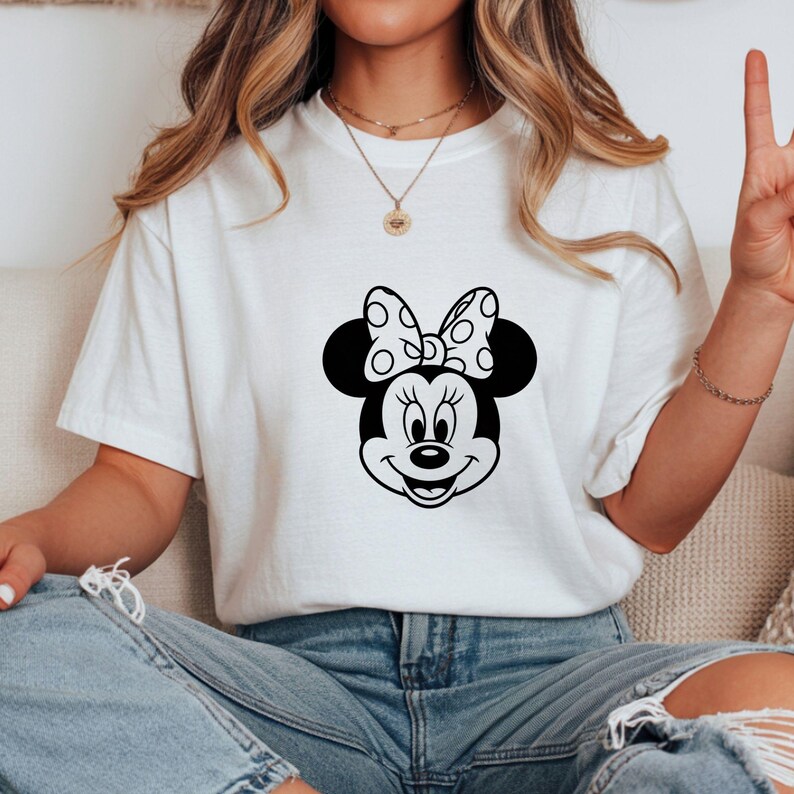 May include: White t-shirt featuring a black and white graphic of Minnie Mouse's face. The design includes her signature polka-dot bow and smiling expression. The shirt is made of a soft material and is suitable for casual wear.