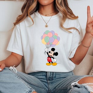 May include: White t-shirt featuring a cartoon Mickey Mouse holding a bunch of pastel-colored balloons. Mickey is depicted in his classic outfit: red shorts, yellow shoes, and black gloves. The balloons are in shades of pink, purple, yellow, and green.