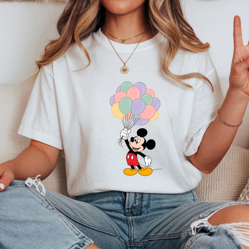 May include: White t-shirt featuring a cartoon Mickey Mouse holding a bunch of pastel-colored balloons. Mickey is depicted in his classic outfit: red shorts, yellow shoes, and black gloves. The balloons are in shades of pink, purple, yellow, and green.