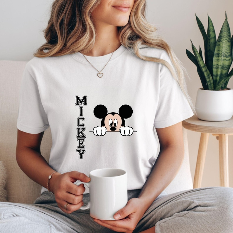 May include: White t-shirt featuring a graphic of Mickey Mouse peeking over a line, with the name "MICKEY" printed vertically in black letters. The person is holding a white mug. The shirt is a casual style.