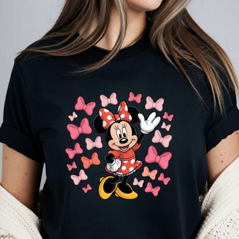 Op de afbeelding: Zwart T-shirt met een cartoonontwerp van Minnie Mouse. Minnie draagt een rode polkadotjurk en strik, omringd door roze strikken. Het personage zwaait met een witte handschoenhand.