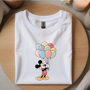 May include: White t-shirt featuring a cartoon Mickey Mouse holding a bunch of colorful balloons. Mickey is wearing red shorts, yellow shoes, and black gloves. The balloons are in shades of pink, blue, green, yellow, and purple.