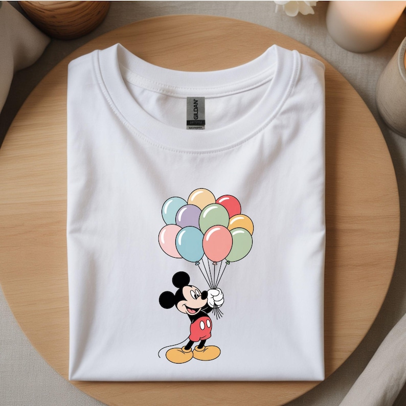 May include: White t-shirt featuring a cartoon Mickey Mouse holding a bunch of colorful balloons. Mickey is wearing red shorts, yellow shoes, and black gloves. The balloons are in shades of pink, blue, green, yellow, and purple.