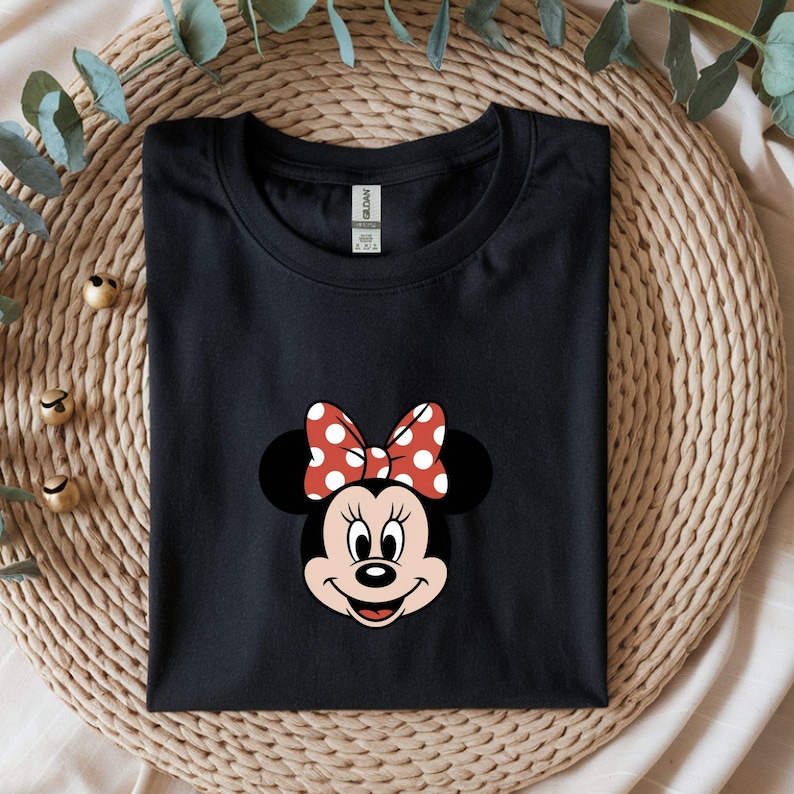 May include: Black t-shirt featuring a Minnie Mouse graphic. The graphic shows Minnie's face with a red bow with white polka dots. The t-shirt is folded and placed on a woven mat.