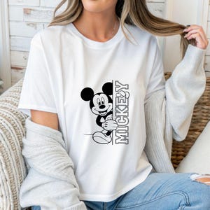 May include: White t-shirt featuring a black and white graphic of Mickey Mouse peeking from behind a vertical text that reads "MICKEY". The cartoon character is smiling. The shirt is a classic crew neck style.