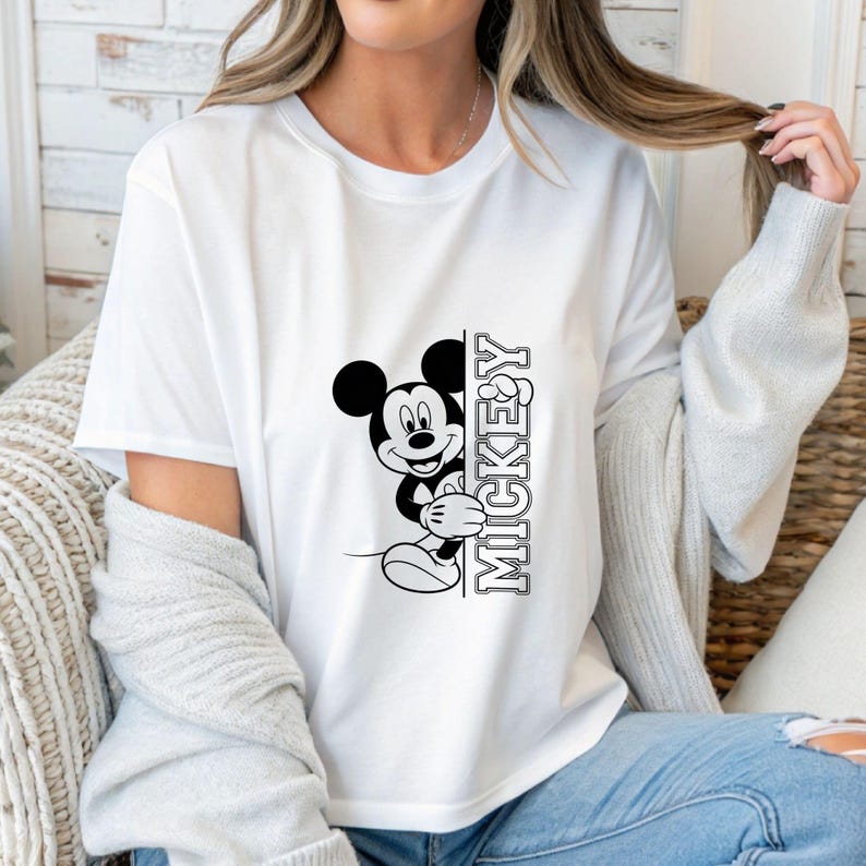 May include: White t-shirt featuring a black and white graphic of Mickey Mouse peeking from behind a vertical text that reads "MICKEY". The cartoon character is smiling. The shirt is a classic crew neck style.