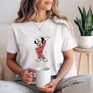 May include: White t-shirt featuring a cartoon Minnie Mouse in a red tracksuit with white accents. Minnie Mouse is wearing a white bow and white sneakers. The t-shirt is a casual style.