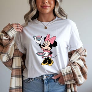 Puede incluir: Camiseta blanca con un dise&ntilde;o de dibujos animados de Minnie Mouse sosteniendo una taza y un platillo. Minnie lleva un vestido y un lazo rosas, orejas negras y zapatos amarillos. Una prenda casual para el d&iacute;a a d&iacute;a.