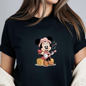 May include: Black t-shirt featuring a cartoon Minnie Mouse playing a pink ukulele while sitting on a hay bale. Minnie Mouse is wearing a pink hat, scarf, and boots. Colorful musical notes float around her.