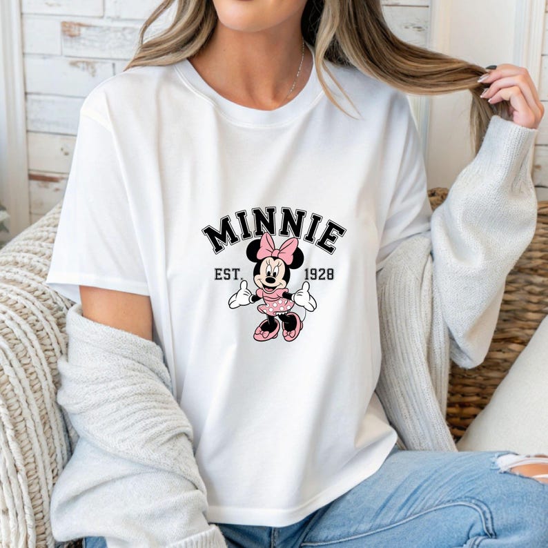 Może przedstawiać: Biała koszulka z grafiką Myszki Minnie z r&oacute;żową kokardą, sukienką i butami. Tekst "MINNIE EST. 1928" jest wydrukowany nad postacią. Projekt jest w kolorach czarnym i r&oacute;żowym.