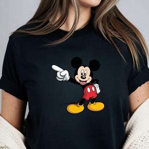 May include: Black t-shirt featuring a cartoon image of Mickey Mouse pointing to the left. Mickey Mouse is depicted with black ears, a white gloved hand, red shorts, and yellow shoes. The shirt is a crew neck style.