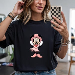 May include: Black t-shirt featuring a cartoon Minnie Mouse dressed as a cowgirl. Minnie Mouse is wearing a pink cowboy hat, dress, and boots. The design is centered on the shirt.