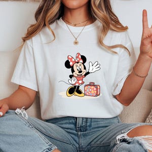 May include: White t-shirt featuring a cartoon Minnie Mouse waving with a suitcase. Minnie Mouse is wearing a red dress with white polka dots and a matching bow. The suitcase is red with travel stickers.