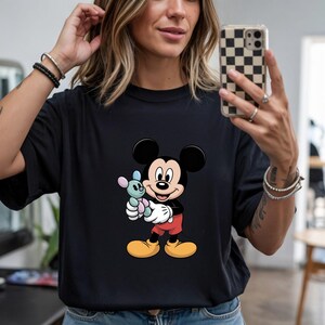 May include: Black t-shirt featuring a cartoon image of Mickey Mouse holding a small stuffed animal. Mickey Mouse is wearing red shorts, yellow shoes, and white gloves. The stuffed animal is blue, pink, and white.