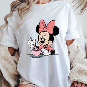 Puede incluir: Camiseta blanca con un dise&ntilde;o de dibujos animados de Minnie Mouse. Minnie lleva un lazo rosa y un vestido rosa, y est&aacute; removiendo una taza de t&eacute; rosa con una cuchara. El dise&ntilde;o est&aacute; centrado en la parte delantera de la camiseta.