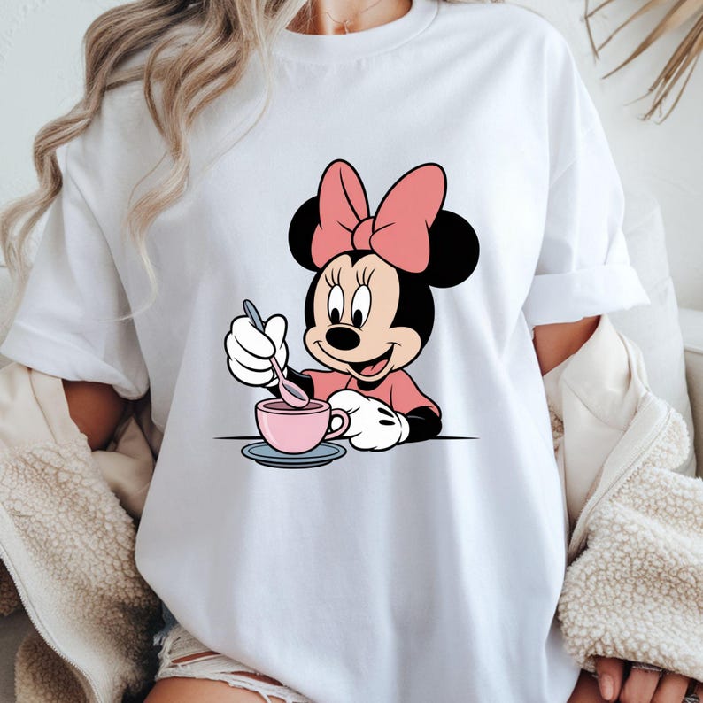 Puede incluir: Camiseta blanca con un dise&ntilde;o de dibujos animados de Minnie Mouse. Minnie lleva un lazo rosa y un vestido rosa, y est&aacute; removiendo una taza de t&eacute; rosa con una cuchara. El dise&ntilde;o est&aacute; centrado en la parte delantera de la camiseta.