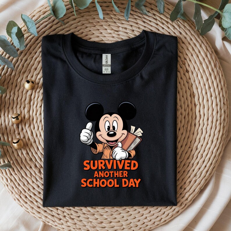 May include: Black t-shirt with a cartoon Mickey Mouse illustration giving a thumbs up and holding a book. The orange text reads "SURVIVED ANOTHER SCHOOL DAY". The shirt is presented on a beige woven mat.