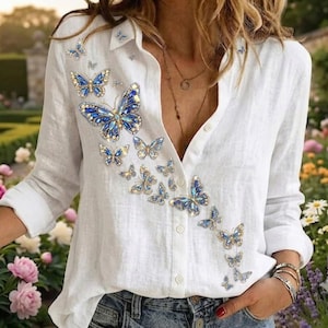 May include: A white long-sleeved button-down shirt featuring a cascading pattern of blue butterfly graphics embellished with sparkling crystals. The shirt has a classic collar, a front button placket, and cuffed sleeves.