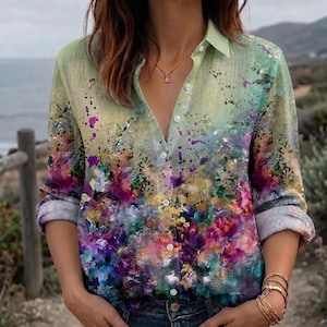May include: A long-sleeve button-down shirt featuring a light green base that transitions into a vibrant, multi-colored abstract paint splatter pattern in shades of purple, pink, blue, and yellow. The shirt has a classic collar and white buttons.