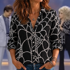 May include: A black long-sleeve button-down shirt featuring a white spiderweb pattern throughout. The shirt has a classic collar, a front button placket with white buttons, and long sleeves with cuffs. The fabric displays a repeating web design.