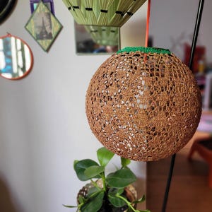 May include: A spherical lampshade featuring a brown, intricate crochet pattern with a green trim at the top. The shade is suspended by an orange cord from a black metal stand, showcasing a textured, open-work design that allows light to pass through.