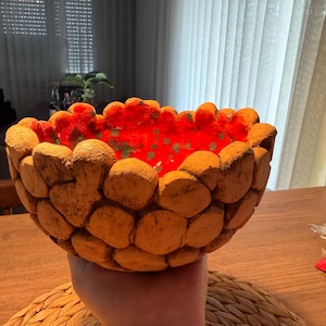 Könnte beinhalten: Eine handgefertigte Schale mit einer strukturierten, steinartigen Außenseite. Die Außenseite der Schale ist warmbraun, während die Innenseite leuchtend rot mit grünen Akzenten ist. Die Schale wird über einer gewebten Matte gehalten.