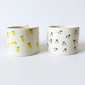 May include: Two off-white ceramic mugs with cylindrical bodies. The left mug features a repeating pattern of yellow lemons with green leaves. The right mug features a repeating pattern of small red cherries with green stems and leaves.