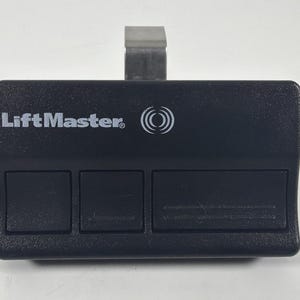Puede incluir: Mando a distancia negro LiftMaster para abridor de puerta de garaje con tres botones y un clip metálico plateado. El logotipo de LiftMaster está impreso en blanco en la parte superior del mando. El mando tiene una superficie texturizada.