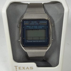 May include: A vintage Texas Instruments digital watch with a silver-tone metal band and case. The watch face is dark blue with white text, including "MULTI ALARM CHRONO DUAL TIME ZONE." The watch is displayed in a white box with the brand name.