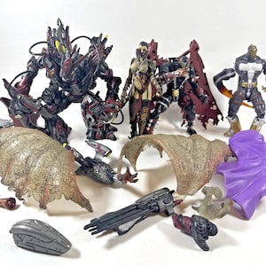 Peut inclure: Plusieurs figurines d'action aux couleurs noir, rouge et gris. Certaines figurines sont équipées de capes et d'armes. Des accessoires tels que des ailes et une cape violette sont également présents. Les figurines sont posées sur une surface blanche.