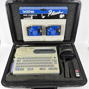May include: A Brother P-touch label maker kit in a black case. The kit includes a beige label maker with a keyboard, a power adapter, and two blue tape cassettes. The case is lined with black foam.