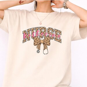 Puede incluir: Camiseta color crema con la palabra "NURSE" en un diseño arqueado y audaz. Las letras tienen un estampado de leopardo con acentos rosas. Un lazo con estampado de leopardo y un gráfico de estetoscopio debajo.