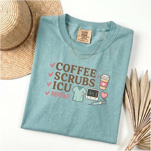 Puede incluir: Una camiseta de color verde azulado claro con las palabras "COFFEE SCRUBS ICU REPEAT" en marrón y rosa. La camiseta presenta ilustraciones de una taza de café, batas, un monitor cardíaco, un corazón y una jeringa. También se ven un sombrero de paja y un collar dorado.
