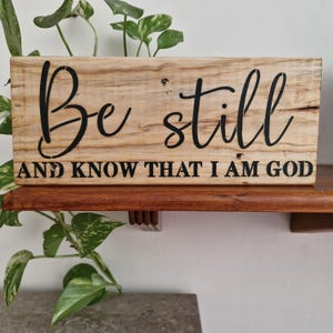 May include: A rectangular wooden sign with the words "Be still and know that I am God" in black script. The sign is made of light-coloured wood. The text is stencilled on the front. The sign is displayed on a wooden shelf.