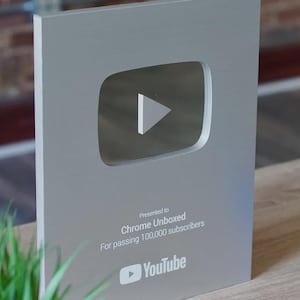 Może przedstawiać: Srebrna nagroda YouTube Play Button, przyznana Chrome Unboxed za przekroczenie 100 000 subskrybentów. Nagroda zawiera ikonę przycisku odtwarzania i logo YouTube. Nagroda jest prezentowana na drewnianym stole.