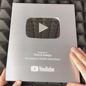 Może przedstawiać: Prostokątna srebrna tabliczka YouTube Creator Award z wyśrodkowaną ikoną przycisku odtwarzania. Tekst brzmi: Presented to Tech & Design For passing 100,000 subscribers, z logo YouTube na dole. Tabliczka ma wykończenie ze szczotkowanego metalu.