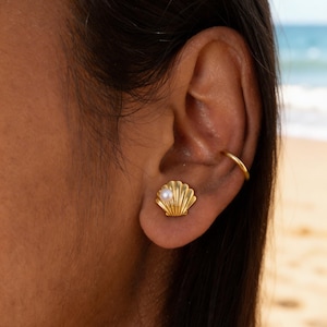Peut inclure: Boucle d'oreille clou en forme de coquillage doré avec une petite perle au centre. La boucle d'oreille est sur l'oreille d'une personne, avec un ear cuff doré visible. L'arrière-plan montre une plage avec l'océan.