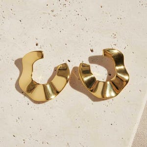 May include: A pair of gold-toned earrings featuring a C-shaped, open-hoop design with a wavy, rippled texture. The earrings have a polished, reflective surface that catches the light, creating distinct highlights and shadows across the curved metal form.
