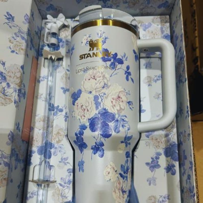 K&ouml;nnte beinhalten: Ein Stanley-Becher mit floralem Muster und gro&szlig;em Griff, mit wei&szlig;en und blauen Rosen auf hellgrauem Hintergrund. Der Becher befindet sich in seiner Originalverpackung, die ebenfalls ein Blumenmuster aufweist. Der Text "Stanley" und "LOVESHACKFANCY" sind sichtbar.