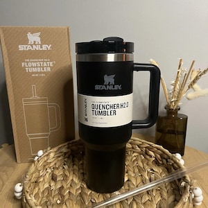 Puede incluir: Un vaso Stanley Quencher H2.0 Flowstate negro con asa, capacidad de 1,18L y pajita. El vaso est&aacute; sobre una bandeja tejida, con la caja al fondo. La caja tiene el logotipo de Stanley y los detalles del producto.