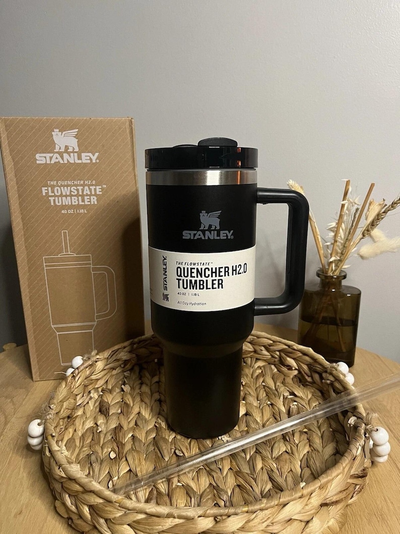 Puede incluir: Un vaso Stanley Quencher H2.0 Flowstate negro con asa, capacidad de 1,18L y pajita. El vaso est&aacute; sobre una bandeja tejida, con la caja al fondo. La caja tiene el logotipo de Stanley y los detalles del producto.