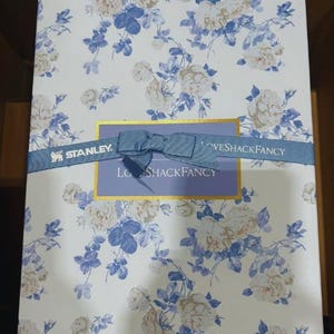 K&ouml;nnte beinhalten: Eine Geschenkbox mit einem floralen Muster in Wei&szlig; und Blau. Ein blaues Band mit einer Schleife umschlie&szlig;t die Box, auf dem die Texte "STANLEY" und "LOVESHACKFANCY" aufgedruckt sind. Die Box hat einen goldenen Rand.
