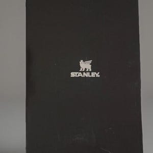Puede incluir: Una caja rectangular negra con el logotipo de Stanley en plata. El logotipo presenta un grifo estilizado con la palabra "STANLEY" debajo. La caja est&aacute; ligeramente inclinada, mostrando sus bordes laterales y superiores.