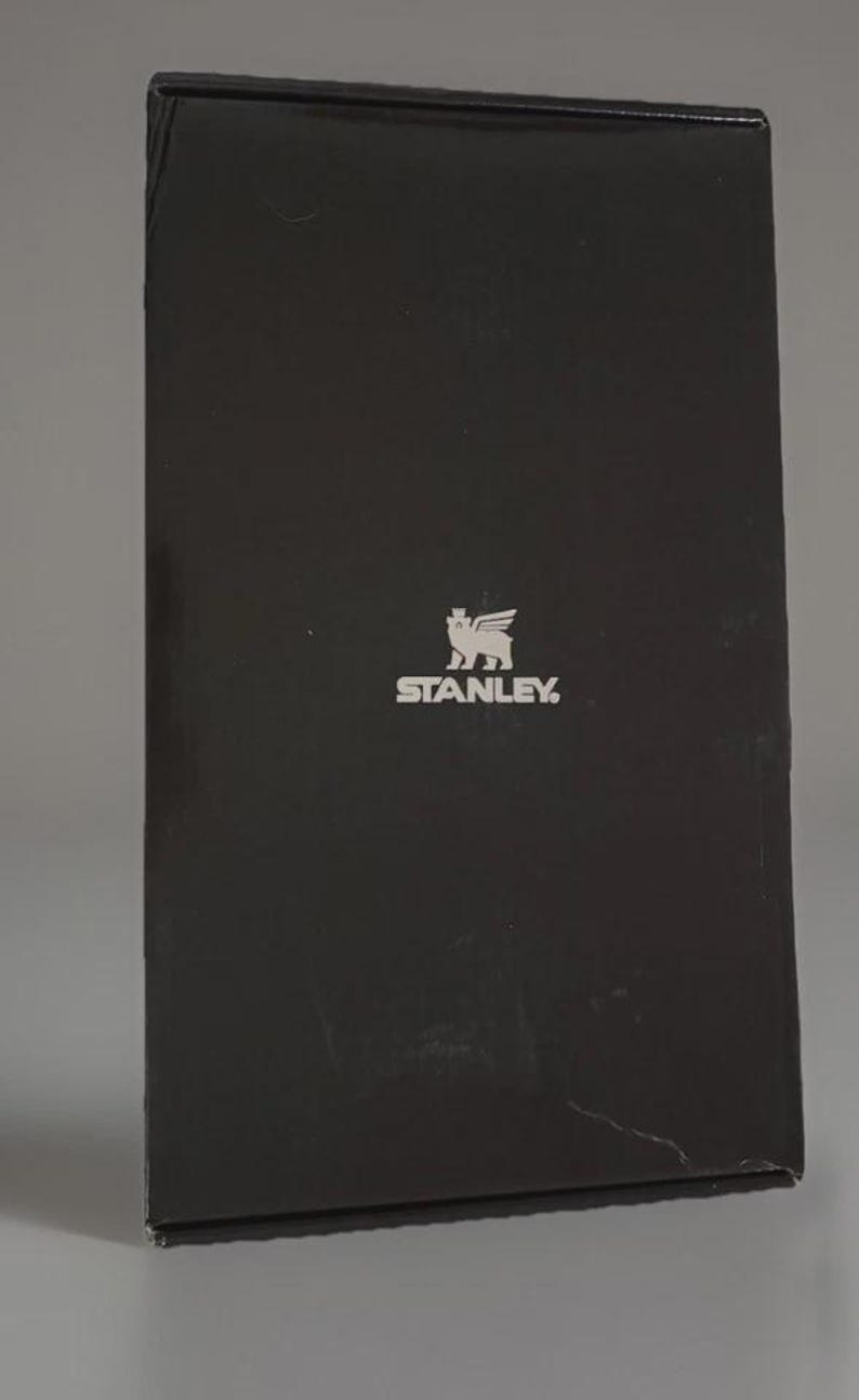 Puede incluir: Una caja rectangular negra con el logotipo de Stanley en plata. El logotipo presenta un grifo estilizado con la palabra "STANLEY" debajo. La caja est&aacute; ligeramente inclinada, mostrando sus bordes laterales y superiores.