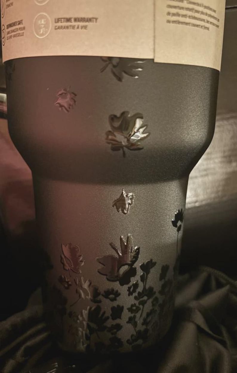 Puede incluir: Un vaso negro con un dise&ntilde;o floral en relieve. Las flores est&aacute;n en relieve sobre la superficie. El vaso tiene una etiqueta de garant&iacute;a de por vida.
