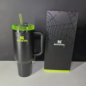 May include: Black Stanley travel mug with a green lid and straw, featuring a spiderweb design. The mug has a handle and the Stanley logo in yellow. A matching black box with a spiderweb design and the Stanley logo is next to the mug.