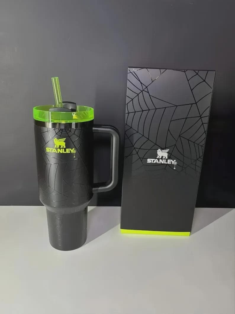 May include: Black Stanley travel mug with a green lid and straw, featuring a spiderweb design. The mug has a handle and the Stanley logo in yellow. A matching black box with a spiderweb design and the Stanley logo is next to the mug.