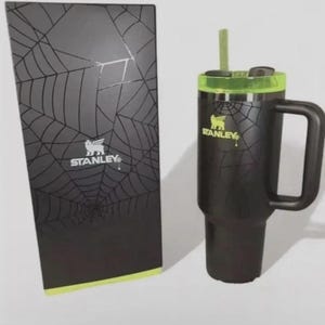 May include: Black Stanley travel mug with a handle, a green rim, and a straw. The mug has a spiderweb design and the Stanley logo. A matching black box with a spiderweb design and the Stanley logo is next to the mug.