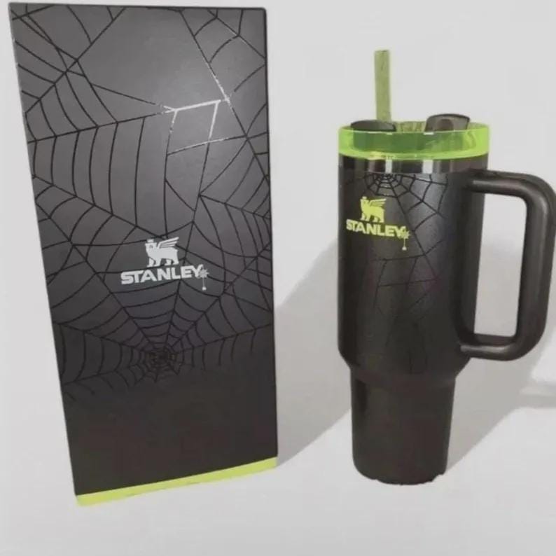 May include: Black Stanley travel mug with a handle, a green rim, and a straw. The mug has a spiderweb design and the Stanley logo. A matching black box with a spiderweb design and the Stanley logo is next to the mug.